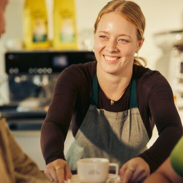 Ein Barista, der in einem lebhaften Café mit einem Lächeln Kaffee serviert und dabei eine warme und einladende Atmosphäre vermittelt.