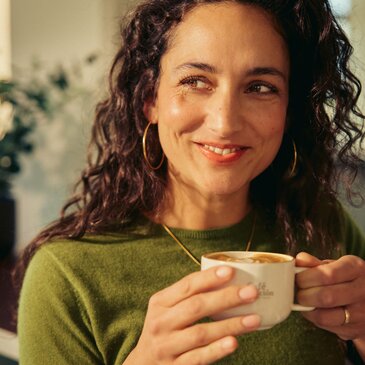 Eine Frau lächelt, während sie in einem gemütlichen Café eine Kaffeetasse in der Hand hält – ein Bild, das eine warme Atmosphäre und ein köstliches Getränk vermittelt.