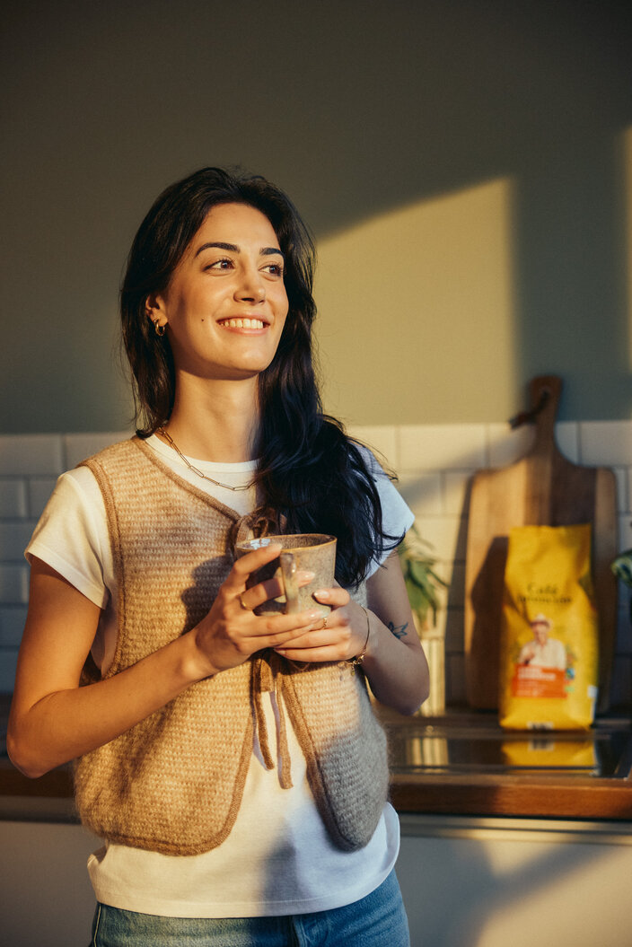 Eine Frau lächelt herzlich, während sie in einer hellen Küche eine Keramiktasse in der Hand hält; im Hintergrund sind Kaffeepackungen zu sehen.
