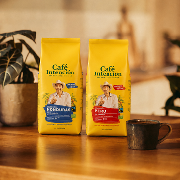 Kaffeepackungen von „Café Intención“ aus Honduras und Peru auf einem Holztisch, im Hintergrund eine Tasse Kaffee und Pflanzen.